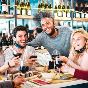 Friends drinking red wine at sushi bar restaurant with open face masks - New normal lifestyle concept with happy people having fun together on vivid filter - Focus on afroamerican guy