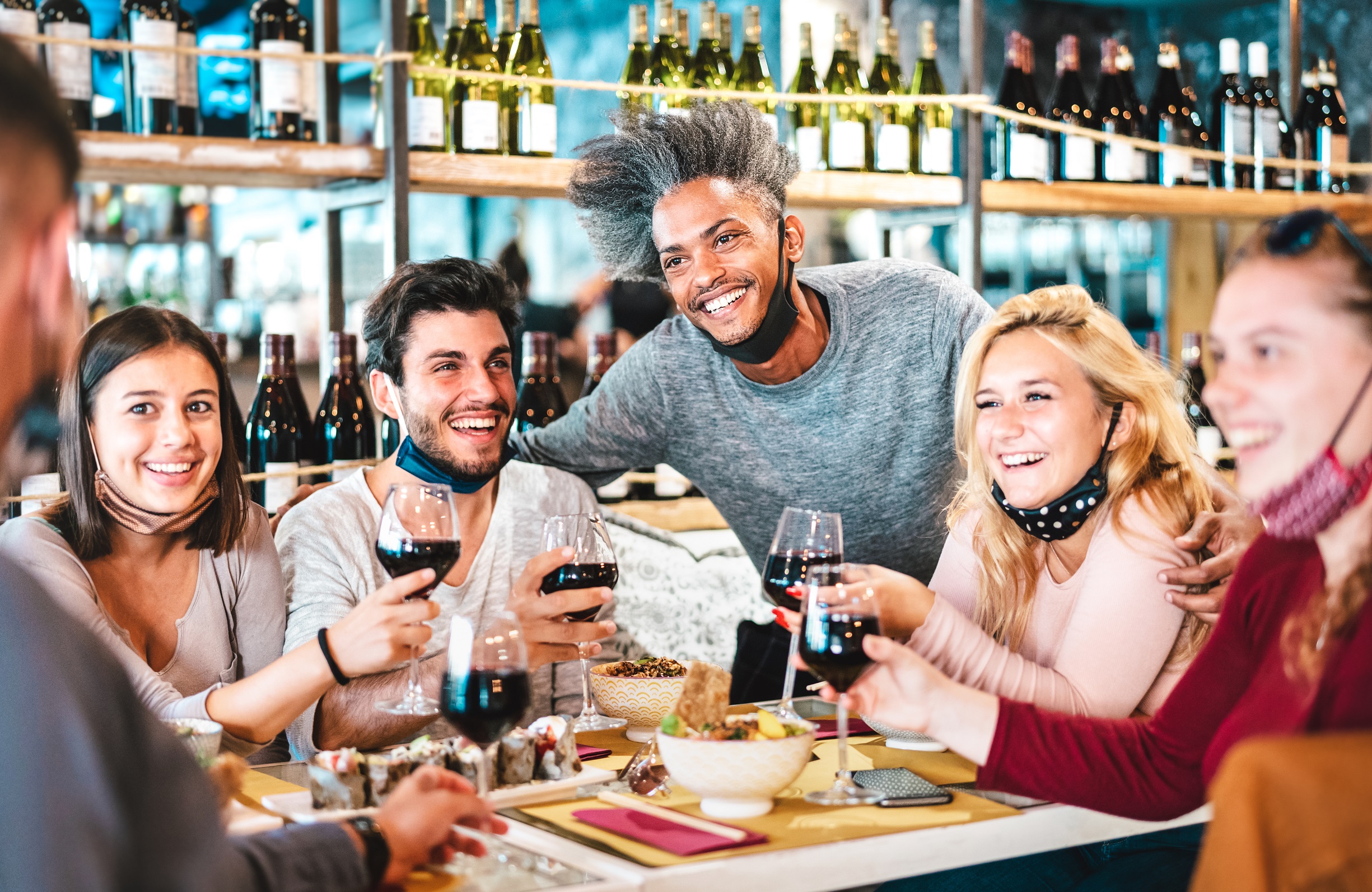 Friends drinking red wine at sushi bar restaurant with open face masks - New normal lifestyle concept with happy people having fun together on vivid filter - Focus on afroamerican guy